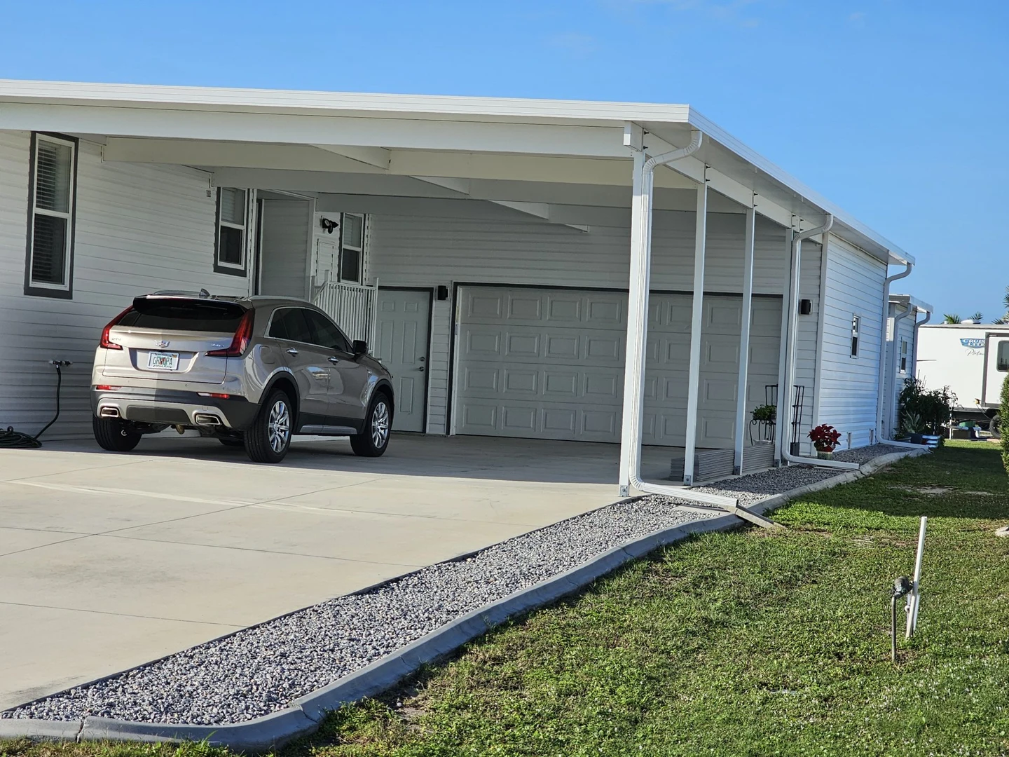 White carport connected to a sided home, with a vehicle beneath the canopy and gravel along the driveway.